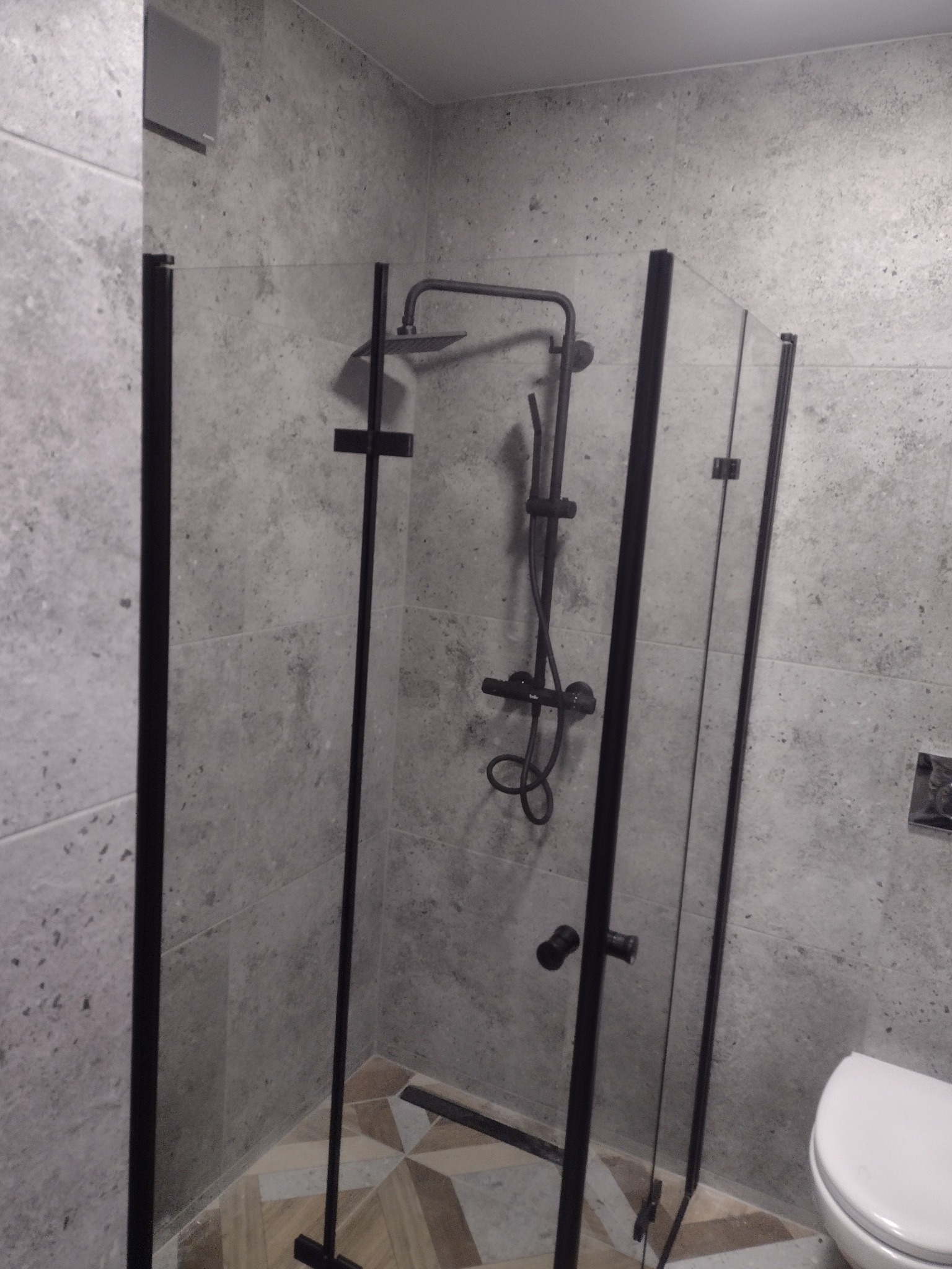 Nowoczesna kabina prysznicowa z czarnymi profilami i deszczownicą, szare płytki imitujące beton, wzorzysta podłoga, widoczny fragment toalety.