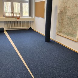 KOMFEX - Niebieska wykładzina dywanowa z jasnym pasem na podłodze w pokoju. Rolka wykładziny oparta o ścianę, okno z widokiem na drzewa w tle.