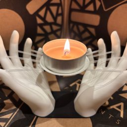 Artur Kowalski - Świecznik z dłoni, wydrukowany w 3D, podtrzymujący zapaloną świeczkę tealight. Dłonie są białe, połączone przezroczystymi gumkami. Tło stanowi wzorzyste pudełko.