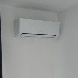 Cool Vibe Sp. z o. o. - Biały klimatyzator ścienny w minimalistycznym wnętrzu, widoczny fragment lampy sufitowej i sofy. Czyste, jasne pomieszczenie.