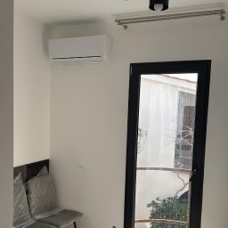 Cool Vibe Sp. z o. o. - Biały klimatyzator na ścianie w jasnym pokoju z czarną lampą sufitową i drzwiami balkonowymi. Dwa krzesła z folią ochronną w rogu. Widok na zewnątrz przez drzwi.