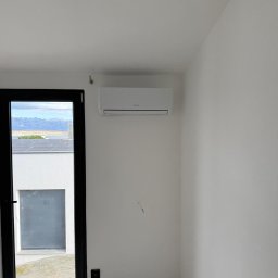 Cool Vibe Sp. z o. o. - Biały klimatyzator na ścianie w jasnym pokoju, widok przez okno na góry i panele słoneczne. Minimalistyczny design, świeżo po montażu.