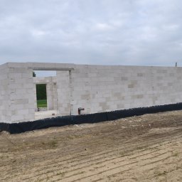 Buduj Z Nami - Budowa domu z białych bloczków: ściany w trakcie wznoszenia, widoczne otwory okienne i drzwiowe, czarna izolacja fundamentów, ziemiste podłoże, pochmurne niebo.