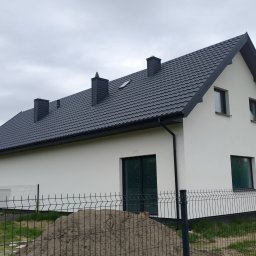 Buduj Z Nami - Nowoczesny dom jednorodzinny z ciemną dachówką i białymi ścianami, widoczny z boku, z nasypem ziemi i ogrodzeniem w tle. Trzy kominy na dachu.