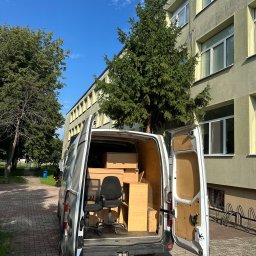 TurboBus - Biały bus dostawczy z otwartymi drzwiami, załadowany czarnym krzesłem biurowym, kartonami i drewnianymi szafkami, stoi na bruku przed jasnozielonym budynkiem.