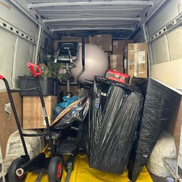 TurboBus - Wnętrze białego busa wypełnione sprzętem fitness (orbitrek, piłka), kartonami i przedmiotami owiniętymi czarną folią, przygotowane do transportu. Na podłodze żółta mata ochronna i wózek.