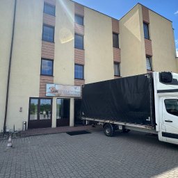 TurboBus - Biały dostawczy truck z czarną plandeką, zaparkowany tyłem do wejścia do beżowego budynku 'SPA & Wellness'. Słońce tworzy refleksy na fasadzie.