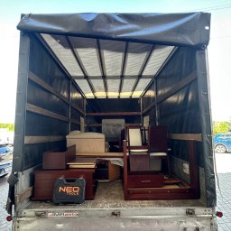 TurboBus - Tył otwartej ciężarówki z plandeką, załadowanej różnorodnymi meblami: krzesłami, beżową sofą, drewnianymi komodami. Na podłodze skrzynka NEO TOOLS. Jasne światło dzienne.