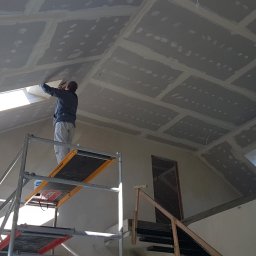 Ciepło Mi - Pracownik na rusztowaniu montuje izolację na poddaszu z oknami dachowymi. Widoczne płyty izolacyjne i szpachlowane łączenia. W tle schody i drzwi.