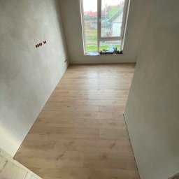 Dominik Samojlik - Jasne panele podłogowe ułożone w pokoju z oknem. Ściany w surowym stanie, widoczne punkty elektryczne. Na parapecie butelki i narzędzia.