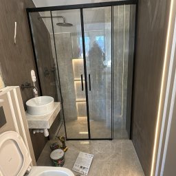MarcinBud - Nowoczesna łazienka w trakcie wykończenia: kabina prysznicowa typu walk-in z czarnymi profilami, umywalka nablatowa i podwieszana toaleta. Widoczne narzędzia i materiały.
