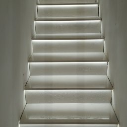 MarcinBud - Białe schody z podświetleniem LED wzdłuż stopni, widok z dołu. Minimalistyczny design, jasne oświetlenie, surowe ściany. Wykończenie wnętrza.