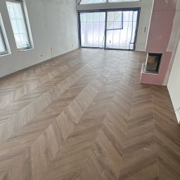 MarcinBud - Ukończona podłoga z parkietu ułożonego w jodełkę w przestronnym, jasnym salonie z dużym oknem i przygotowanym miejscem na kominek. Widoczne zabezpieczenia okien folią.