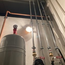 DMT INSTAL - Zainstalowany system grzewczy w Gorzowie Wielkopolskim, z widocznymi miedzianymi i ocynkowanymi rurami, zbiornikiem wyrównawczym i dwoma pompami obiegowymi, zamocowany na jasnej ścianie.