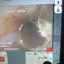 DMT INSTAL - Ekran z inspekcji kamerą wewnątrz rury, ukazujący zatory i korzenie. Widoczne dane techniczne, operator trzymający panel sterowania.