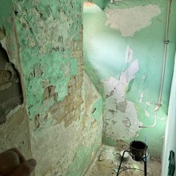 DMT INSTAL - Pomieszczenie w trakcie remontu z łuszczącą się zieloną farbą na ścianach, odsłaniającą tynk i cegły, z widocznymi rurami i gruzem na podłodze.