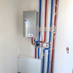Usługi Hydrauliczne RAWEX - Zmontowany piec gazowy z instalacją: pionowe rury w kolorach niebieskim i czerwonym, biała szafka pod piecem. Widok na ścianę w pomieszczeniu technicznym.