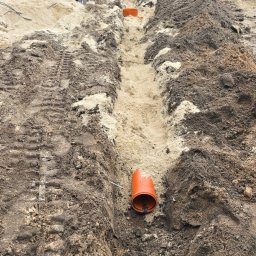 Usługi Hydrauliczne RAWEX - Wykopany rów pod instalację z widocznymi pomarańczowymi rurami kanalizacyjnymi. W tle dom jednorodzinny i ogrodzenie. Widoczne ślady opon koparki.