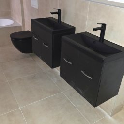 Usługi Hydrauliczne RAWEX - Wykończona łazienka z dwoma czarnymi umywalkami z szafkami, czarną toaletą i beżowymi płytkami. Widoczne białe rurki na ścianie.