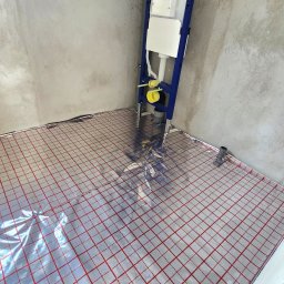 Usługi Hydrauliczne RAWEX - Mata grzewcza rozłożona na podłodze w surowym pomieszczeniu, z widocznym stelażem podtynkowym WC. Instalacja w trakcie realizacji, przed wylaniem posadzki.