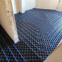 Usługi Hydrauliczne RAWEX - Niebieskie rurki ogrzewania podłogowego ułożone na czarnej macie w surowym wnętrzu. Widok z góry na system instalacji przed wylewką.