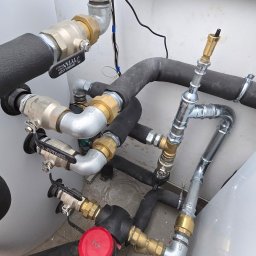 Usługi Hydrauliczne RAWEX - Złożona instalacja hydrauliczna z rurami w izolacji, zaworami i licznikiem z czerwoną obudową. Widoczne złącza mosiężne i fragment buta instalatora w dolnym rogu.