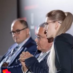 Yana Shevchuk - Panel dyskusyjny z udziałem trzech osób: dwóch mężczyzn w garniturach i kobiety z mikrofonem, prawdopodobnie tłumaczki, podczas konferencji.