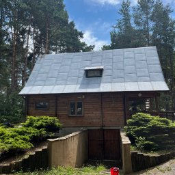 Dach-Cat Jacek Gąsienica - Drewniany dom z szarym dachem z blachy trapezowej, otoczony zielenią drzew i krzewów. Widoczny betonowy zjazd do piwnicy i czerwone wiadro.