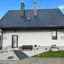 Dach-Cat Jacek Gąsienica - Dom jednorodzinny z ciemnoszarą dachówką i jasną elewacją. Widoczna lina, prawdopodobnie do prac na wysokości. Uwagę zwracają dwa kominy i zadbany ogród.