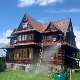 Dach-Cat Jacek Gąsienica - Czyszczenie elewacji drewnianego domu z dachem z blachy na wysokości. Pracownik w kasku używa myjki ciśnieniowej. Widoczna drabina i sprzęt do mycia.