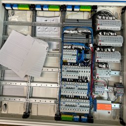Sinus Energy - Rozdzielnica elektryczna w trakcie montażu, widoczne przewody, zabezpieczenia i moduły KNX. Na obudowie leżą dokumenty z opisem instalacji.