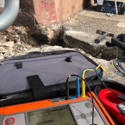 Sinus Energy - Miernik rezystancji uziemień w trakcie pomiaru przy rurze spustowej. Widoczne kable pomiarowe, torba narzędziowa i fragment wykopu.