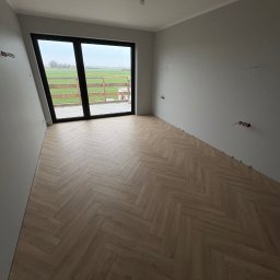 Benbud - Jasny parkiet ułożony we wzór jodełki w nowym, pustym pokoju z dużym oknem balkonowym i szarymi ścianami. Widoczne wystające kable elektryczne.