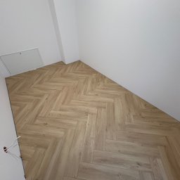 Benbud - Jasny parkiet ułożony we wzór jodełki francuskiej w rogu pomieszczenia. Widoczna klapka rewizyjna w ścianie i wystające przewody elektryczne.