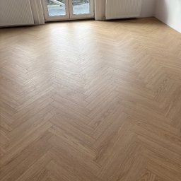 Benbud - Jasny parkiet ułożony we wzór jodełki francuskiej w przestronnym pokoju z białymi ścianami, grzejnikami i oknem balkonowym. Widok na zieleń za oknem.