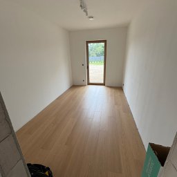 Benbud - Jasne panele podłogowe w nowym pomieszczeniu z białymi ścianami, widok na zewnątrz przez drewniane drzwi. Torba narzędziowa i karton w rogu kadru.