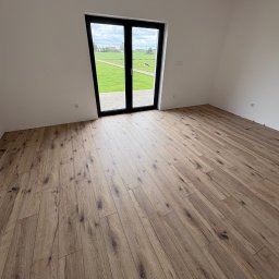 Benbud - Ułożone panele podłogowe w jasnym pokoju z widokiem na zielone pole przez czarne, przeszklone drzwi balkonowe. Naturalne światło podkreśla wzór drewna.
