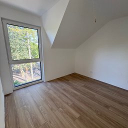 Benbud - Jasne panele podłogowe w pokoju na poddaszu z oknem. Widok na zieleń za oknem. Białe ściany i sufit. Wystający kabel z sufitu. Nowe wykończenie wnętrza.