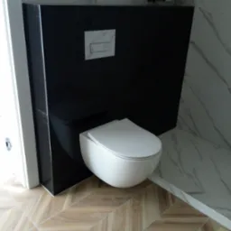 Nowoczesna toaleta podwieszana z czarną zabudową, biały przycisk spłukujący, płytki imitujące marmur na ścianach i drewnopodobna podłoga ułożona w jodełkę.