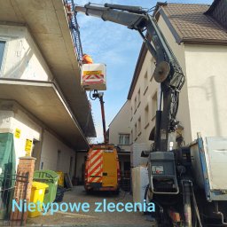 BMT - Pracownik w podnośniku koszowym, na tle budynku, wykonuje prace na wysokości. Widoczny samochód z podnośnikiem i fragment otoczenia.