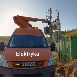 BMT - Pomarańczowy samochód Iveco z podnośnikiem koszowym na tle słupa energetycznego i kontenera. Prace elektryczne w terenie, oświetlenie uliczne.