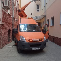 BMT - Pomarańczowy podnośnik koszowy Iveco na brukowanym dziedzińcu, ramię podnośnika sięga do klimatyzatora na ścianie budynku. Otwarta boczna furtka.