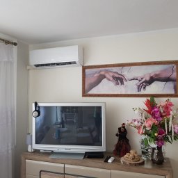 Bartłomiej Figas Klimatyzacja - Biała klimatyzacja ścienna w salonie, nad telewizorem i obrazem 'Stworzenie Adama'. Kwiaty w wazonie i dekoracje na komodzie. Zasłony w oknie po lewej.