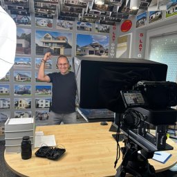 FMD.studio - Uśmiechnięty mężczyzna w okularach, z uniesioną ręką, stoi za biurkiem z kamerą. W tle ściana z fotografiami domów i oświetlenie studyjne.