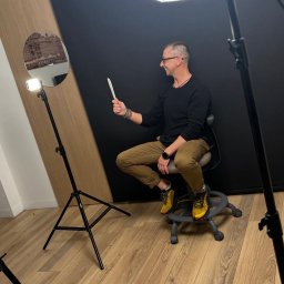 FMD.studio - Mężczyzna w okularach siedzi na krześle w studio fotograficznym, trzymając w dłoni biały reflektor. W tle czarne tło i lampa pierścieniowa z odbiciem miasta.
