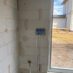 Amperio Instalacje Elektryczne - Niebieskie puszki elektryczne z przewodami na ścianie z betonu komórkowego w surowym stanie, widok z wnętrza budynku w trakcie budowy.