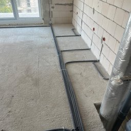 Amperio Instalacje Elektryczne - Szare rury instalacji elektrycznej ułożone na surowym betonie w nowym budynku. Widoczne puszki elektryczne w ścianie z białych bloczków. Okno w tle.