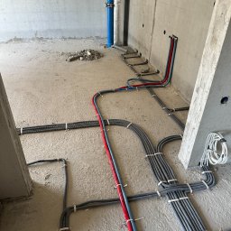 Amperio Instalacje Elektryczne - Szare rury w karbowanej osłonie, przymocowane do podłoża w surowym stanie, prowadzone wzdłuż ściany. Dodatkowo widoczne rury w kolorze czerwonym i niebieskim.