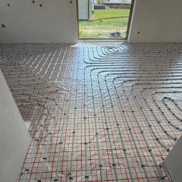 Dan-Kan Instalacje Grzewcze I Sanitarne Daniel Kania - Białe rury ogrzewania podłogowego ułożone spiralnie na folii z czerwoną siatką w jasnym pomieszczeniu z oknem na zewnątrz. Widok z góry, etap instalacji.