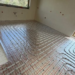Dan-Kan Instalacje Grzewcze I Sanitarne Daniel Kania - Ogrzewanie podłogowe w Niedźwiadzie: białe rury ułożone na folii z czerwoną siatką w surowym wnętrzu. Widok na okno i krajobraz za nim.